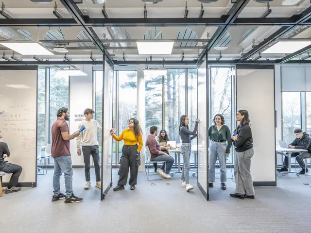 Wide shot of the Maths Gym, with students standing working on different problems on large whiteboards.