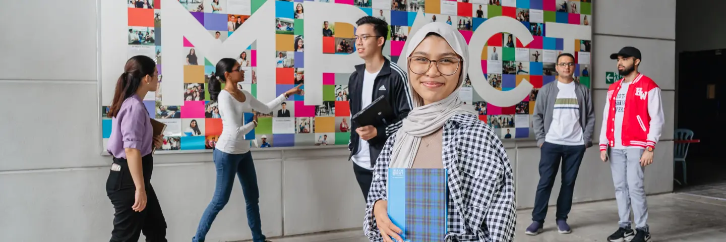 A woman stands in front of a colourful mural, people walking behind her as she smiles holding a book.