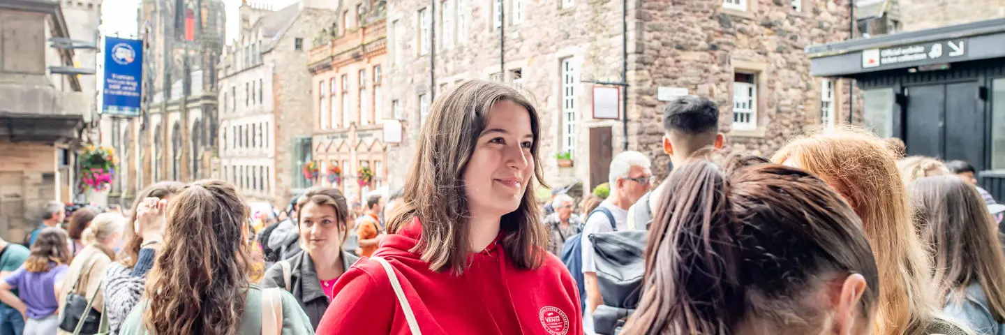Student wearing a red hoodie and holding a canvas bag smiles in conversation.